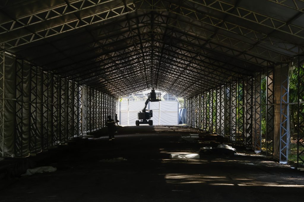 Fotografia mostra o interior de uma tenda montada com estrutura metálica. O espaço está iluminado pela luz natural que vem do lado direito, ao fundo da imagem. 