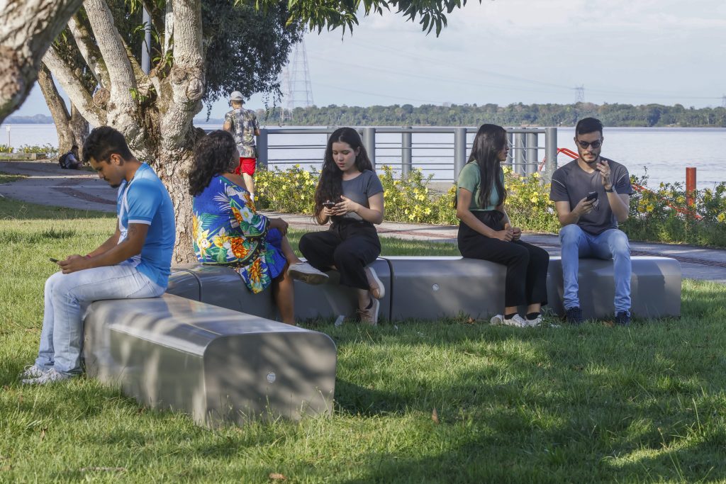 Fotografia de dia, próximo à orla do rio Guamá, em destaque, um banco comprido, em formato curvo. Algumas pessoas estão sentadas no banco. 