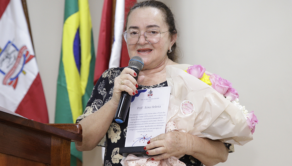 Fotografia da professora Rosa Helena. Ema é uma mulher branca, de cabelos escuros e usa óculos de armação de metal no formato quadrado. A professora segura um microfone com a mão direita e um buquê de flores rosa e uma placa com seu noma na mão esquerda.