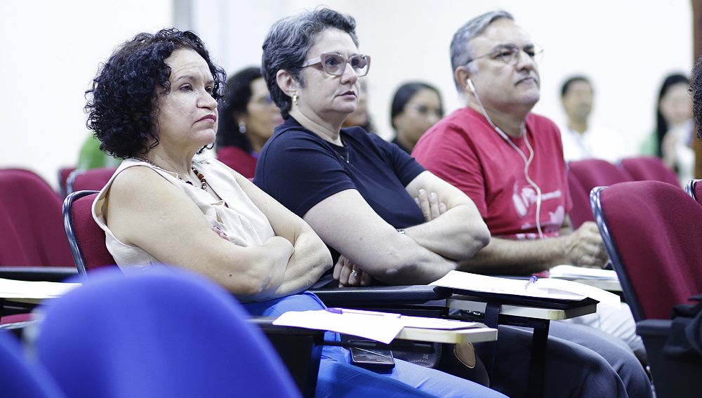 Fotografia de parte de uma plateia. Na foto, em destaque, estão três pessoas, um homem e duas mulheres que olham na mesma direção.