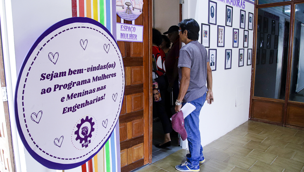 Fotografia mostra uma placa em primeiro plano escrito “Sejam bem-vindas(os) ao Programa Mulheres e Meninas nas Engenharias”. Ao fundo, algumas pessoas estão dentro da sala, enquanto outras passam pela porta. Na porta está uma placa escrito “Espaço mulher”.