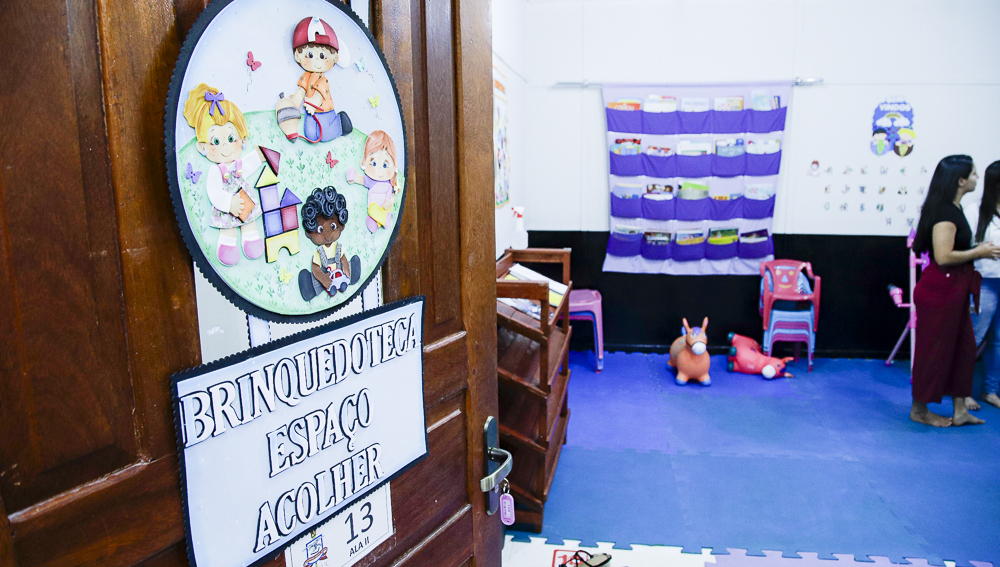 Fotografia mostra uma porta entreaberta com uma placa pendurada, com a seguinte escrita: Brinquedoteca espaço acolher. Ao fundo estão duas pessoas, pequenas cadeiras e alguns brinquedos. O chão da sala é coberto por tatames azuis.