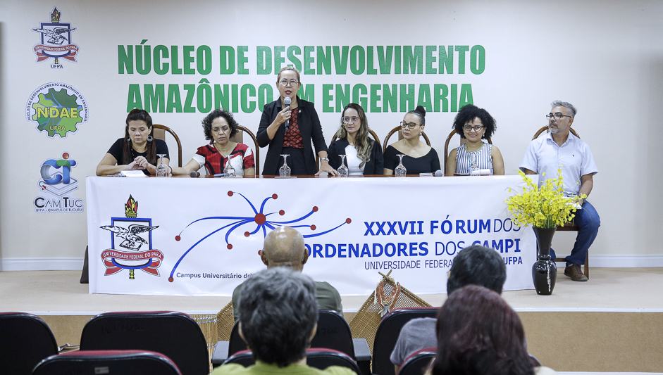 Fotografia registra a mesa de um evento. Nela há seis mulheres e um homem. Ao centro, uma mulher fala ao microfone, enquanto é observadas pelos outros integrantes. Ela veste blazer e calça pretos e uma blusa vermelha.