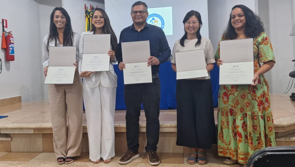 Foto posada mostra quatro mulheres e um homem, dispostos em pé em uma escada, segurando placas nas mãos. Todos sorriem. Ao fundo, uma tela é projetada por trás de uma mesa com pano azul.