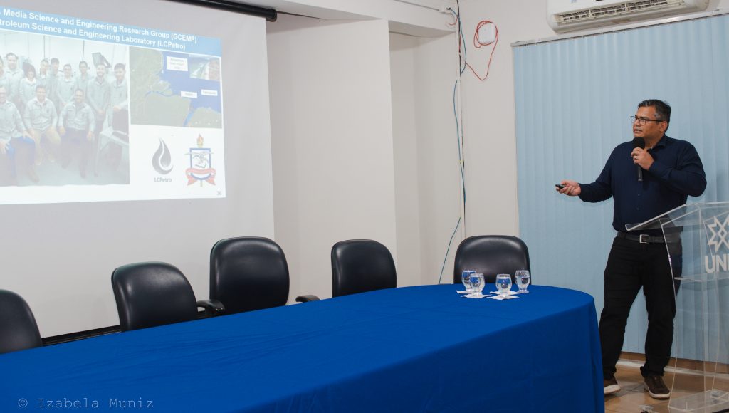 Fotografia do professor Pedro Tupã Pandava Aum. Ele veste uma blusa de manga comprida na cor azul marinho e calça preta. O professor está em pé falando ao microfone e apontando para uma projeção em uma tela na parede, que mostra fotos, mapas e o brasão da UFPA. Em frente à tela de projeção está uma mesa com um pano azul sobre ela.