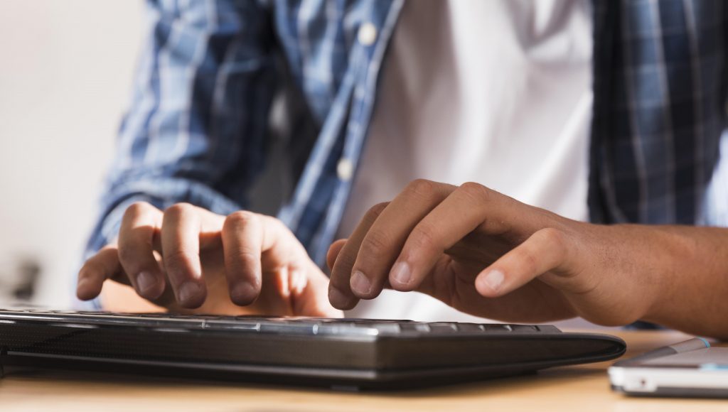 Fotografia, em plano fechado, de mãos masculinas digitando em um teclado de computador.