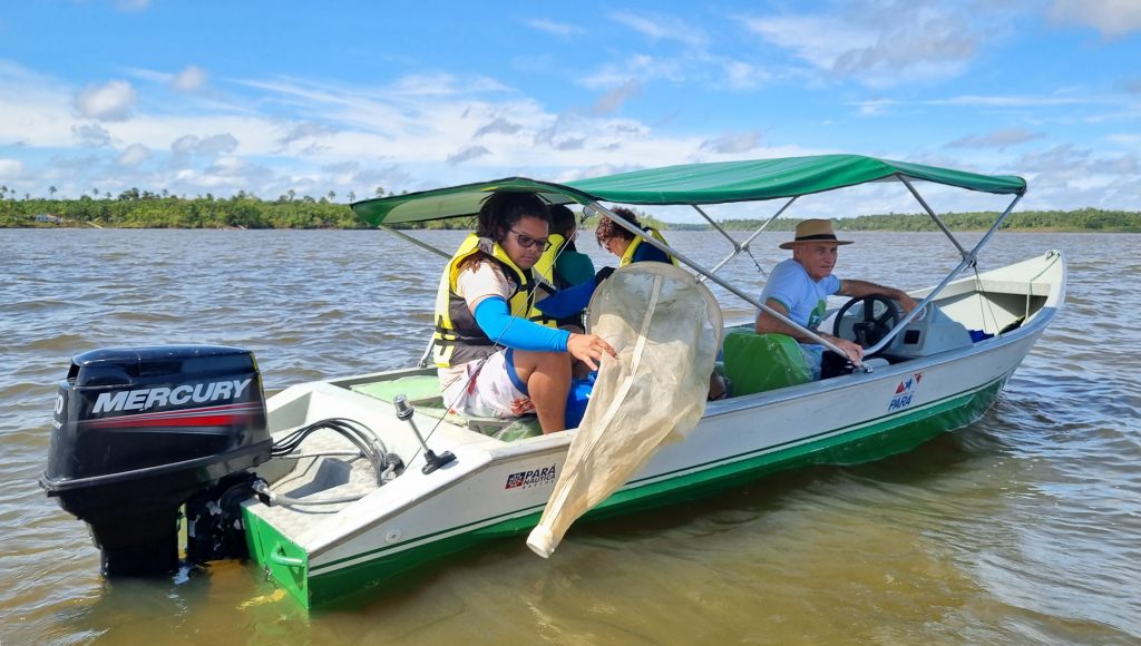 Fotografia mostra uma pequena embarcação no meio de um rio. Dentro da embarcação há quatro pessoas, sendo uma delas em destaque, na lateral da embarcação. Essa pessoa segura uma rede de pesca, em formato de funil, pendurada em um suporte.circular.