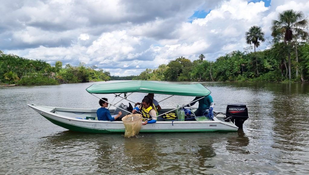 Fotografia mostra uma pequena embarcação no meio de um rio. A paisagem é composta por várias árvores às margens desse rio. Dentro da embarcação há quatro pessoas. Uma das pessoas segura uma rede de pesca, pendurada em um suporte circular.