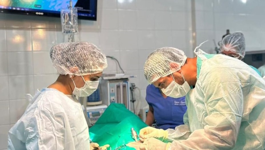 Fotografia retrata uma sala de cirurgia. No centro da imagem, estão uma mulher e um homem olhando para um tecido verde entre eles. Eles estão vestindo, cada um, uma roupa hospitalar descartável, uma touca sanfonada e uma máscara de cor branca.