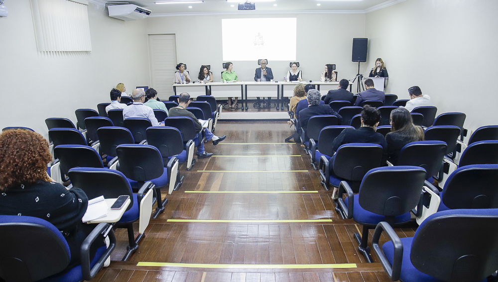 Fotografia de um auditório com cadeiras azuis. Parte da plateia do auditório está ocupada. No fundo da imagem, há uma mesa com cinco pessoas sentadas. No canto direito da imagem, uma mulher está atrás de um púlpito.
