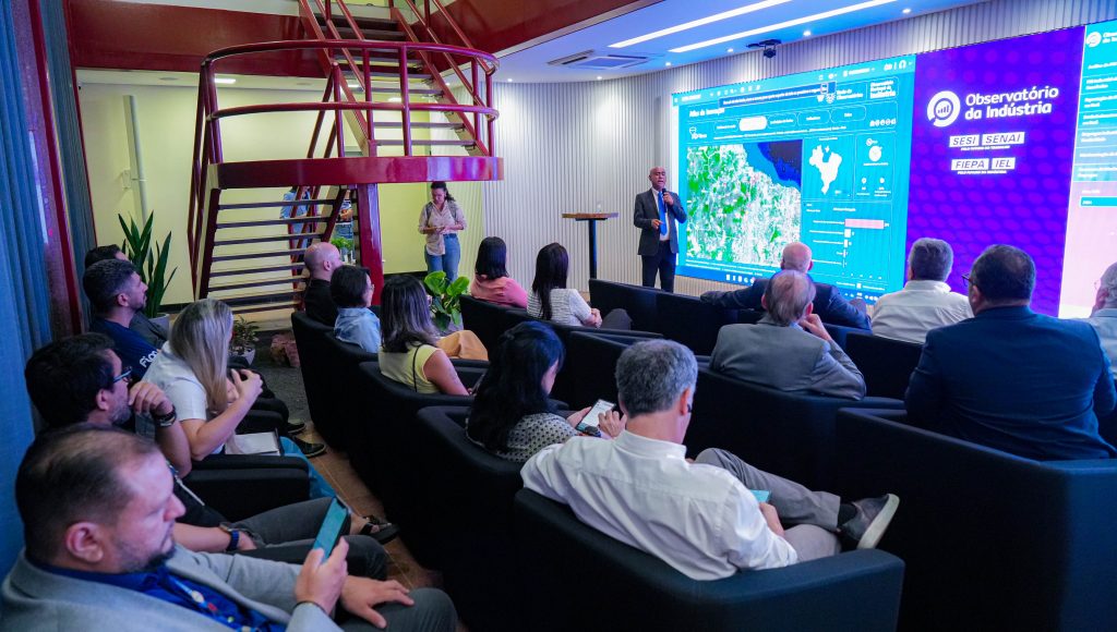 Fotografia registra uma pequena plateia que observa o reitor da UFPA falando ao microfone. Ao fundo, uma grande imagem é projetada. è possível ver o título: OBSERVATÓRIO DA INDÚSTRIA.