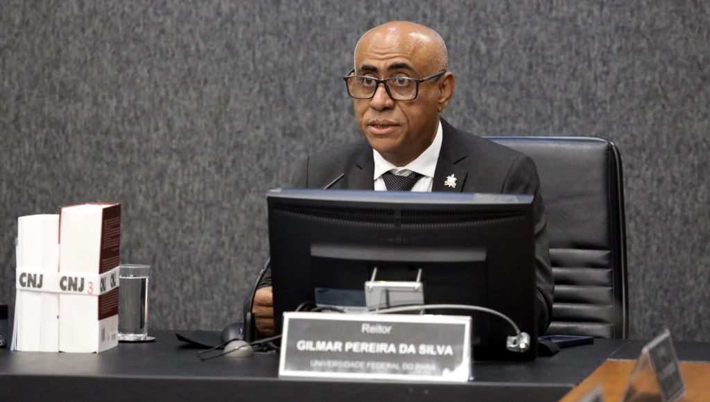 Fotografia do reitor da UFPA, Gilmar Pereira da Silva, falando em um microfone acoplado a uma mesa. A frente dele há um tela que se assemelha com tela de computador.