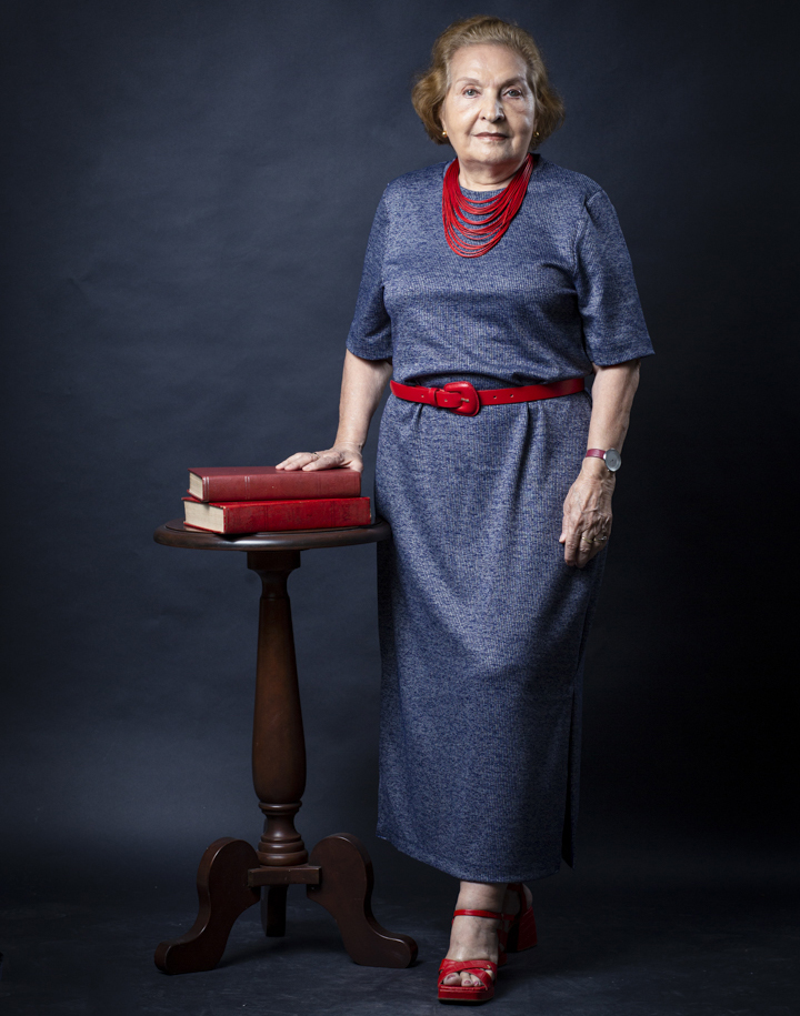 Fotografia posada da professora Violeta Loureiro. Ela é uma mulher branca, de cabelos curto castanho claro, usa um vestido azul com cinto e colar vermelhos. Ela usa sapatos vermelhos e sorri. Ao lado da professora, há uma mesa de madeira com dois livros vermelhos. O fundo da foto é escuro, em tons que variam do preto ao cinza mais escuro.