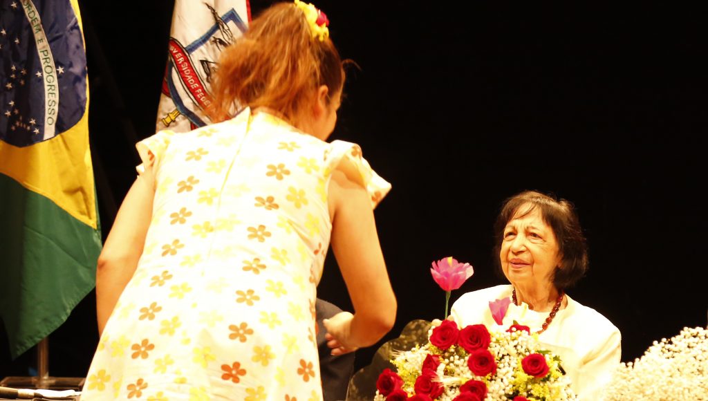 Fotografia de Maria Sylvia Nunes sendo presenteada com uma flor. A sua frentes está uma mulher de vestido florido. Ao fundo da foto, é possível identificar a bandeira da UFPA e a do Brasil.