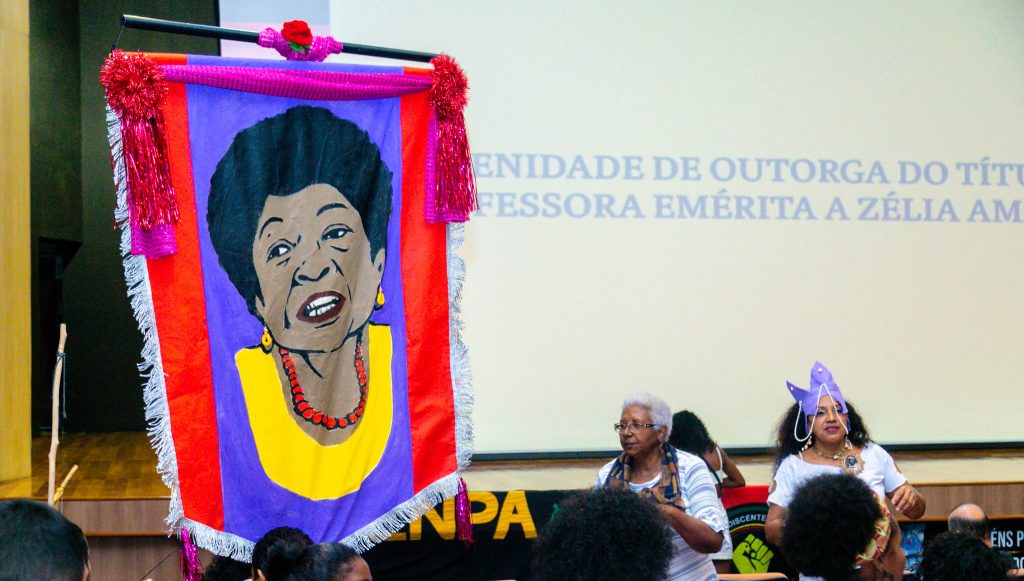 Fotografia registra um estandarte que reproduz a imagem da professora Zélia Amador de Deus. O estandarte é composto por tecidos nas cores vermelho e roxa, com franjas brancas. Ao fundo da imagem, é possível ler em um telão: Outorga do título de professora emérita à Zélia Amador