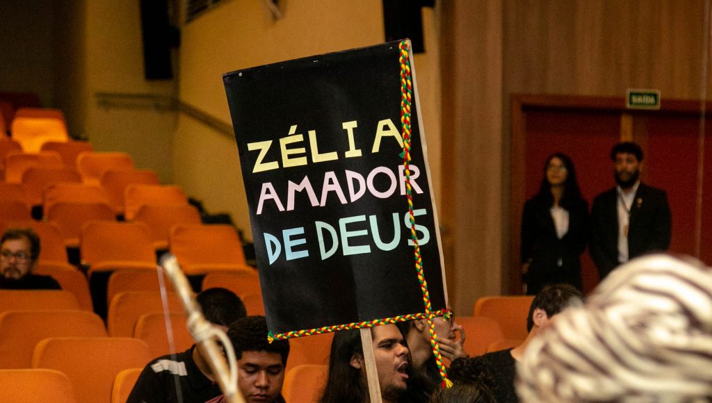 Fotografia de um cartaz com o texto: ZÉLIA AMADOR DE DEUAS. O cartaz tem fundo preto com letras coloridas.