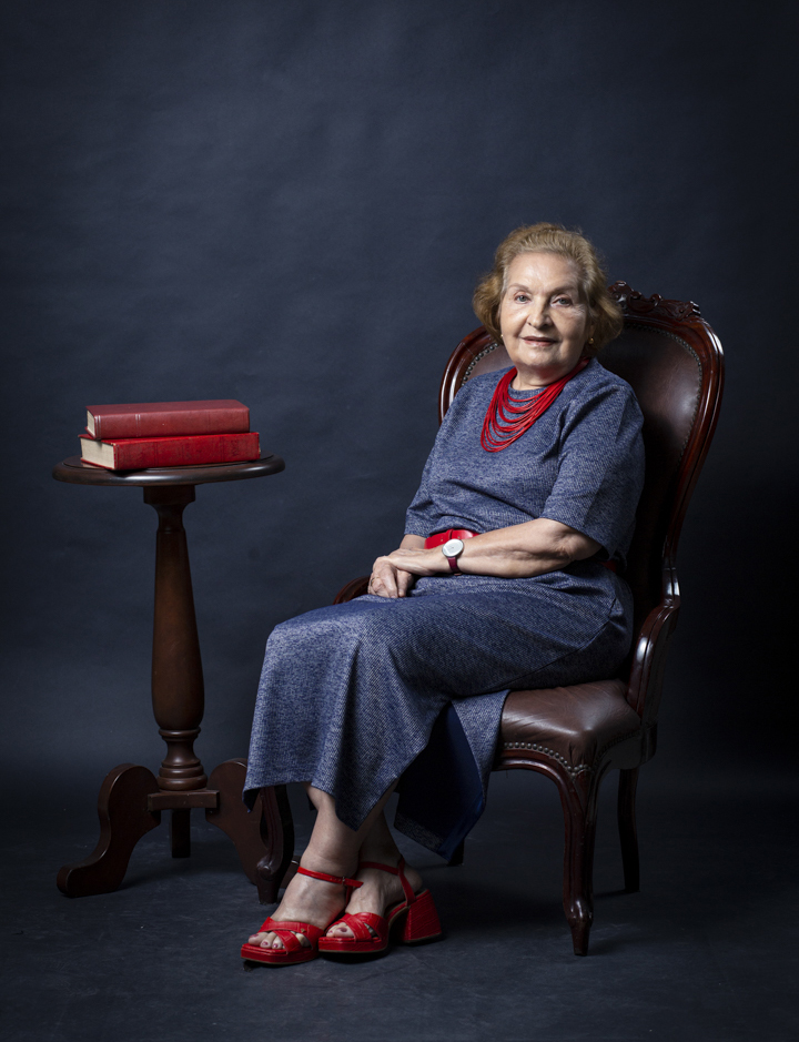 Fotografia posada da professora Violeta Loureiro. Ela é uma mulher branca, de cabelos curto castanho claro, usa um vestido azul com cinto e colar vermelhos. Ela usa sapatos vermelhos e está sentada em uma poltrona de madeira e estufado marrom. Ao lado da professora, há uma mesa de madeira com dois livros vermelhos.