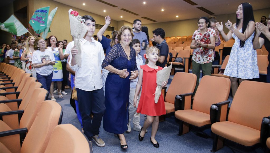 Fotografia do cortejo que abriu a cerimônia de outorga do título de professora Emérita à Ana Tancredi. A professora está a frente do cortejo, de mãos dadas com duas crianças. Um menino e uma menina. As crianças seguram buquês de flores.