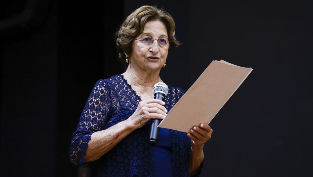 Fotografia da professora Ana Tancredi falando ao microfone. Ela segura folhas de papel. 