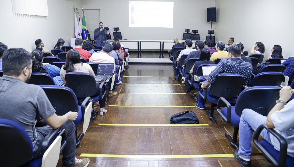 Fotografia de um auditório. A foto registra o público sentado em poltronas azuis, em primeiro plano. Ao fundo, é possível identificar um home que fala ao microfone e um grande tela com imagens projetadas.