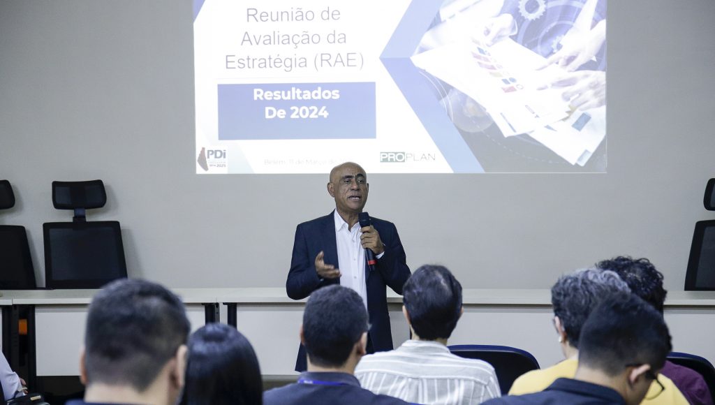 Fotografia registra o reitor da UFPA, Gilmar Pereira da Silva, falando ao microfone durante uma apresentação. Ao fundo, há um telão que projeta a seguinte informação: Reunião de Avaliação da Estratégia (RAE), resultados de 2024.