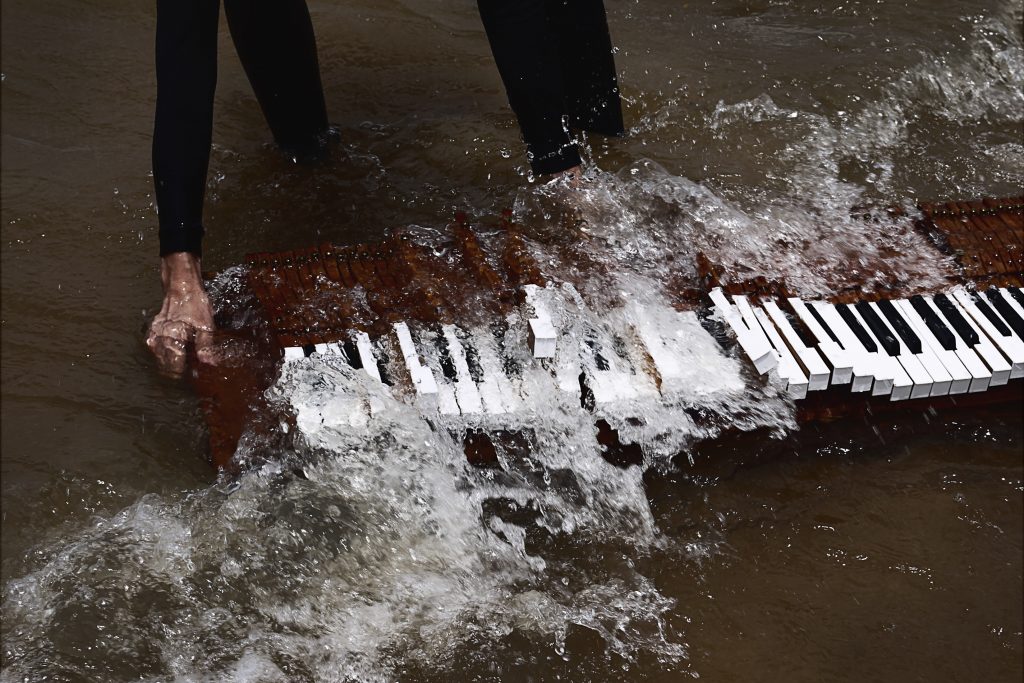 Um teclado de piano aos pés de uma pessoa. Ambos estão dentro de um rio. Foto Danielle Fonseca.