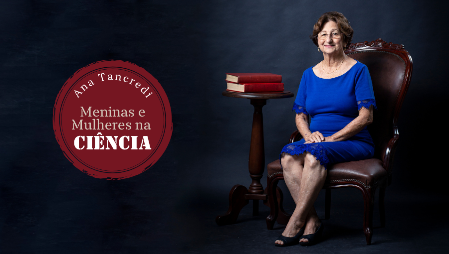 Fotografia da professora emérita Ana Tancredi. Ela está sentada em uma cadeira de madeira, ao lado de uma mesa de madeira com dois livros de capa vermelha. A professora é branca, tem cabelo curto liso e veste um vestido azul. Ela está com as mãos cruzadas sobre as pernas. Sobre a foto um selo com o seguinte texto: Ana Tancredi, MENINAS E MULHERES NA CIÊNCIA.