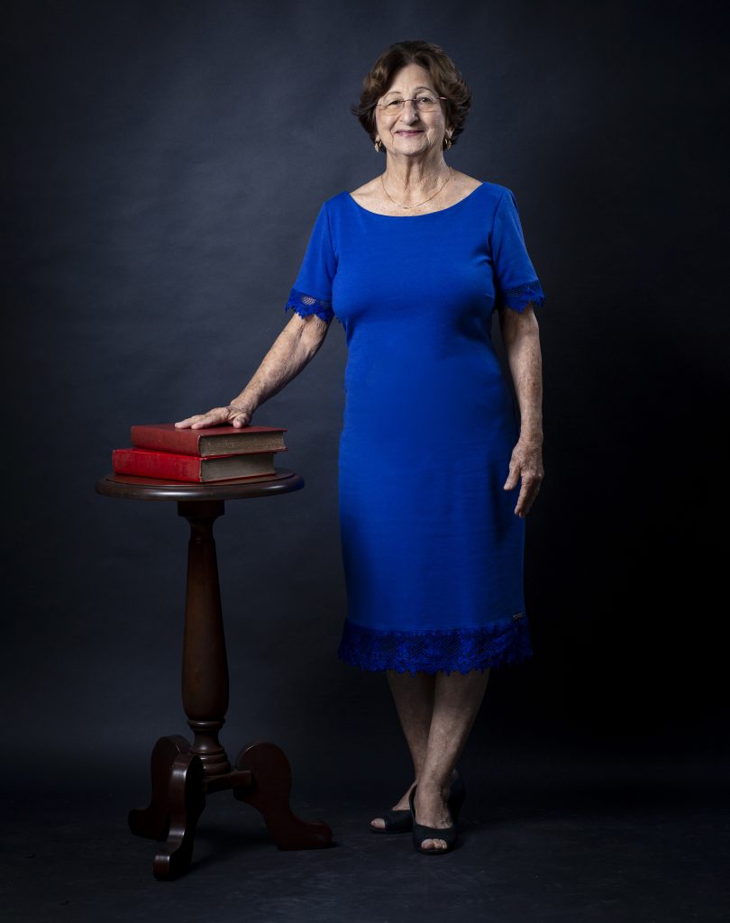 Fotografia da professora emérita Ana Tancredi. Ela está em pé ao lado de uma mesa de madeira com dois livros de capa vermelha. A professora é branca, tem cabelo curto liso e veste um vestido azul.