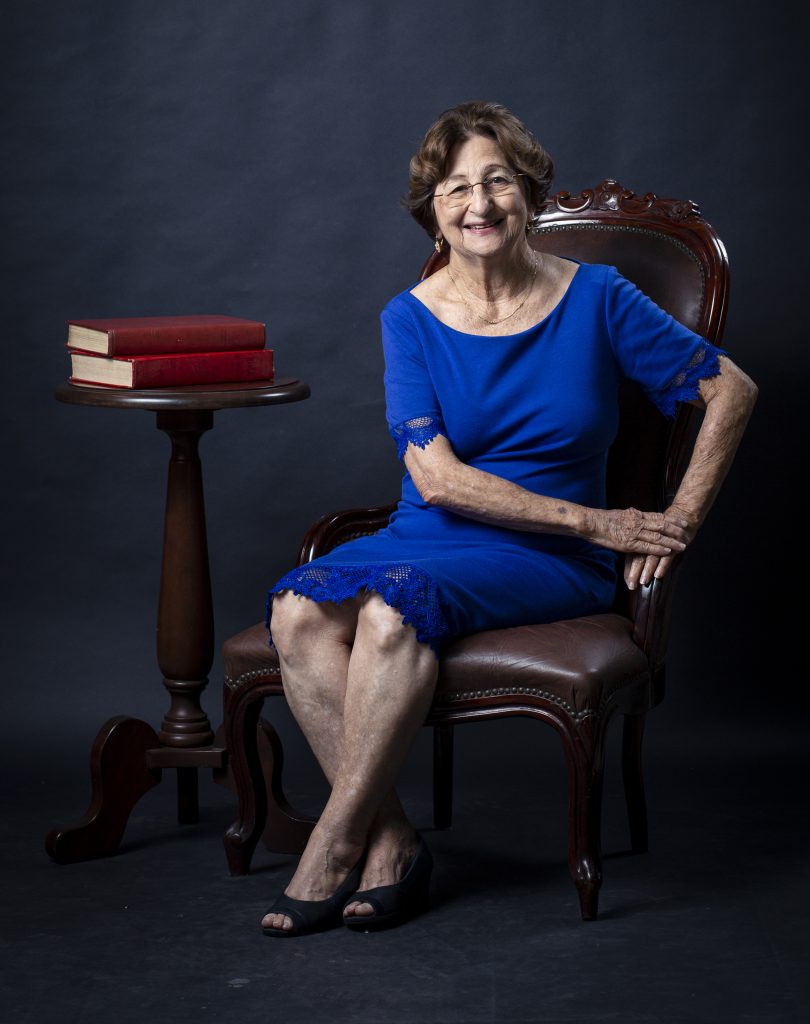 Fotografia da professora emérita Ana Tancredi. Ela está sentada em uma cadeira de madeira, ao lado de uma mesa de madeira com dois livros de capa vermelha. A professora é branca, tem cabelo curto liso e veste um vestido azul. Ela está com as mãos sobre o braço da cadeira.