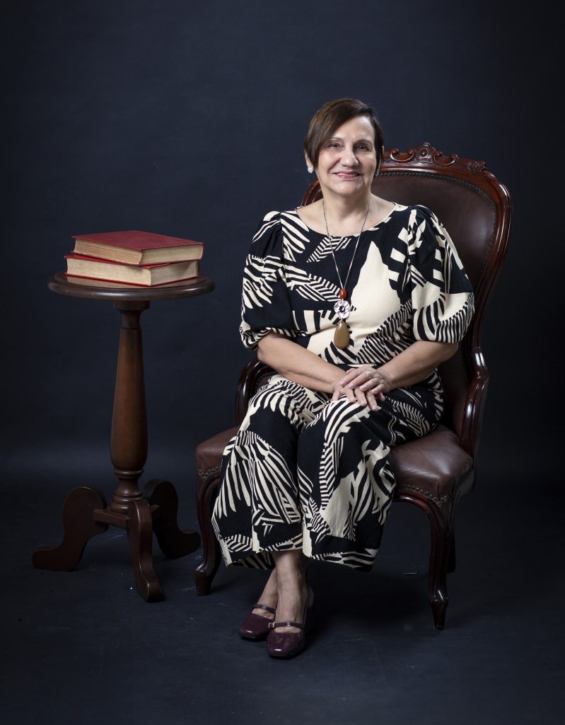 Fotografia posada de Maria de Nazaré Sarges. Ela está sentada em uma cadeira de madeira com estofado marrom. Veste um macacão nas cores preta e bege. Ela é uma mulher parda, de cabelos curtos, lisos na altura do ombro.