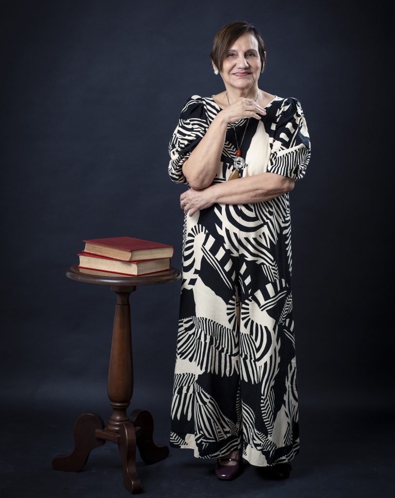 Fotografia posada de Maria de Nazaré Sarges. Ela está EM PÉ, AO LADO DE UMA MESA COM DOIS LIVROS DE CAPAS VERMELHAS. Veste um macacão nas cores preta e bege. Ela é uma mulher parda, de cabelos curtos, lisos na altura do ombro.