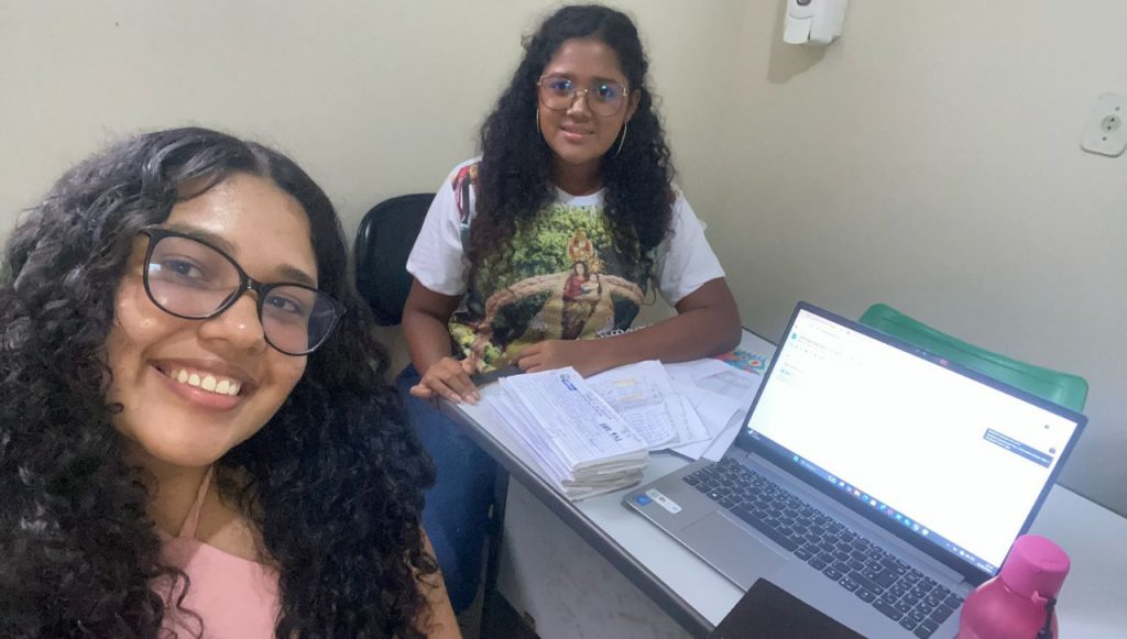 Fotografia de duas meninas, elas olham para nós e sorriem. Uma delas está em primeiro plano e a outra atrás. Elas estão sentadas em cadeiras em volta de uma mesa com um notebook e papéis em cima. As duas usam óculos e têm cabelos cacheados.