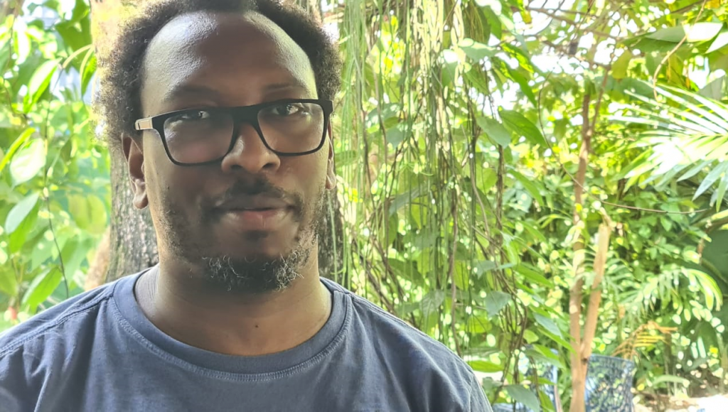 Fotografia posada de Mamadou Ba. Ele é um homem negro, de cabelos curtos, barba e bigode. 
