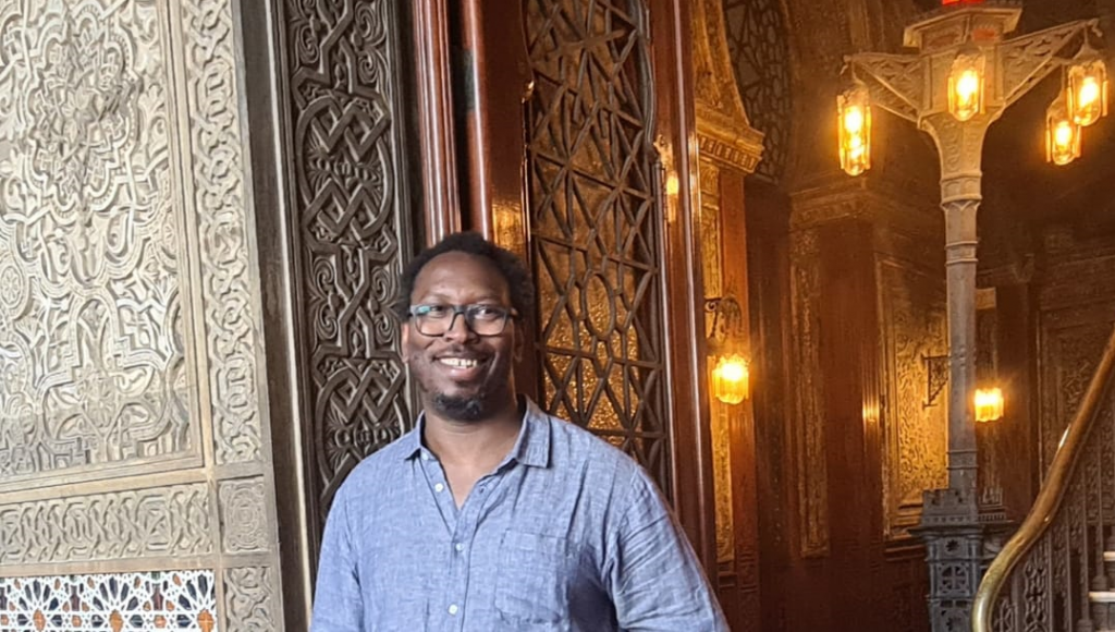 Fotografia posada de Mamadou Ba. Ele é um homem negro, de cabelos curtos, barba e bigode. Ele usa óculos de armação preta quadrada e uma blusa azul de manga comprida.