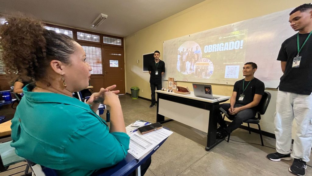fOTOGRAFIA REGISTRA PARCIALMENTE UMA SALA DE AULA. nA IMAGEM, TRÊS HOMENS DE PRETO ESTÃO OBSERVANDO UMA MULHER DE VERDE FALAR. aTRÁS DOS HOMENS, HÁ UMA PROJEÇÃO EM UM QUADRO BRANCO QUE MOSTRA A PALAVRA "OBRIGADO"
