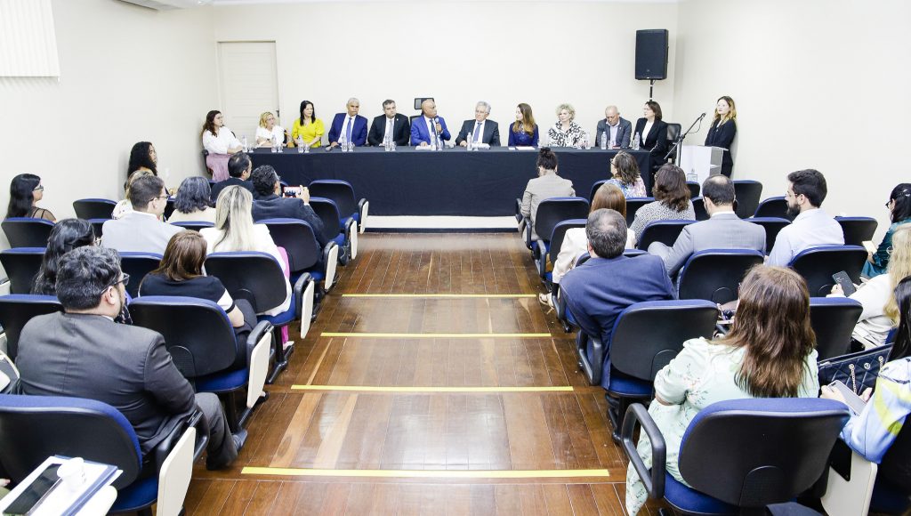 Fotografia de um auditório durante um evento. Na imagem é possível observar uma mesa ao fundo, onde um grupo de pessoas está sentado.