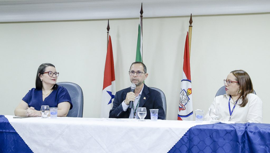 Fotografia da mesa de um evento. nela estão três pessoas, duas mulheres e um homem. Atrás deles há três mastros com as bandeiras do Pará, do Brasil e da UFOA, nesta ordem.