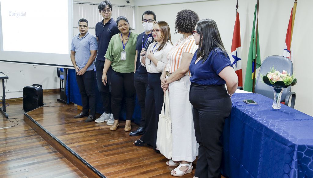 Fotografia de um grupo de sete pessoas, três homens e quatro mulheres. Uma das mulheres seguram um microfone. Eles estão em pé em frente a uma mesa e ao lado de um telão.