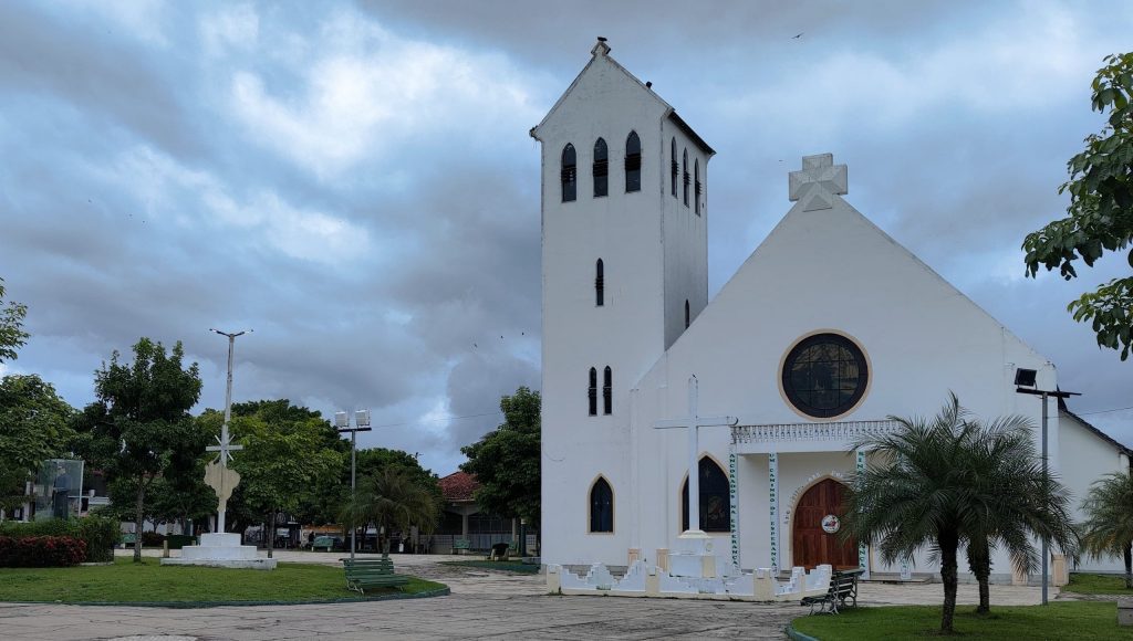 Fotografia da fachada de uma igreja. Ela é toda branca, composta por uma torre e porta de madeira.