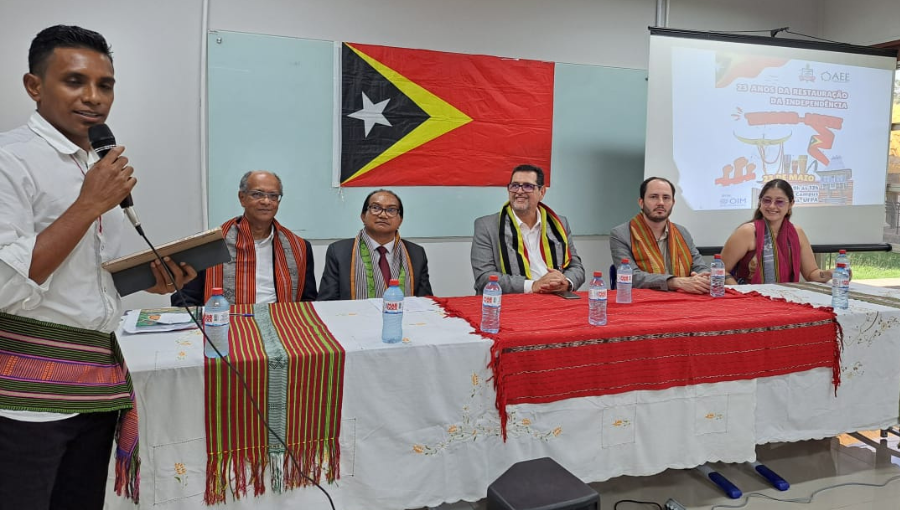 Fotografia de um grupo de pessoa na mesa de um evento. Atrás da mesa, cinco pessoas estão sentadas. Na lateral da mesa, um homem fala ao microfone. Ao fundo da imagem, é possível identificar uma bandeira do Timor Leste pendurada na parede.