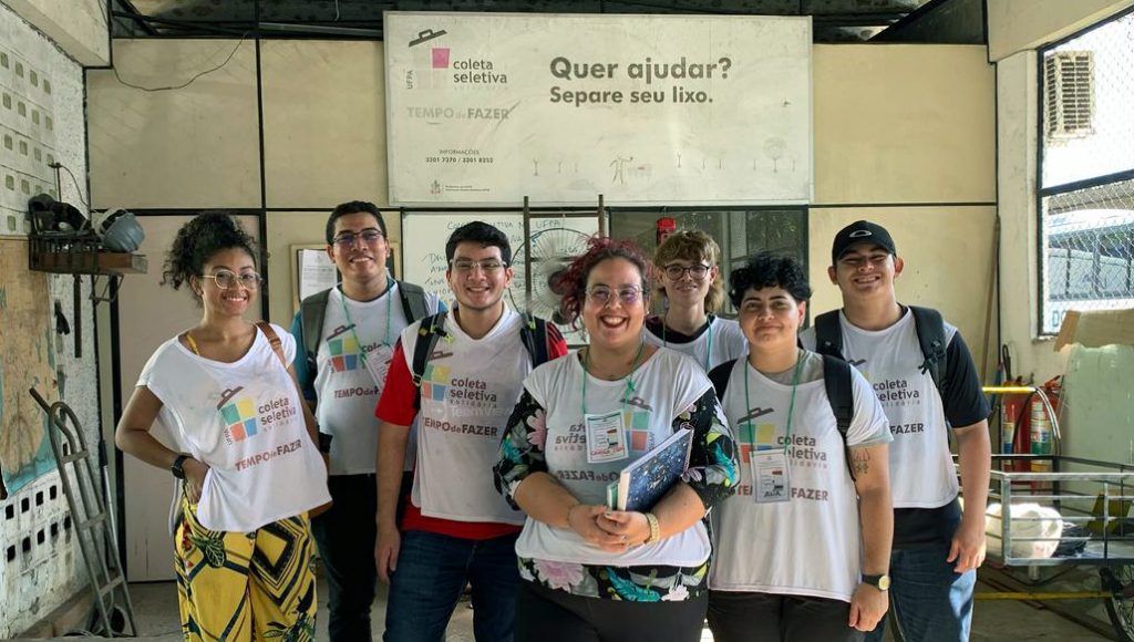 Fotografia de um grupo de jovens vestido com coletes brancos com a seguinte identificação: COLETA SELETIVA. Os jovens estão a porta de uma sala, que tem um cartaz "Quer ajudar? Separe o seu lixo".