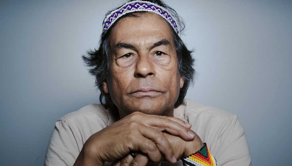 Fotografia posada de Ailton Krenak, um homem indígena, de pele parda e cabelos curtos pretos. Ele este apoiando o seu rosto em suas mãos. Usa uma adereço na cabela e uma pulseira no braço esquerdo, ambos de miçangas coloridas.