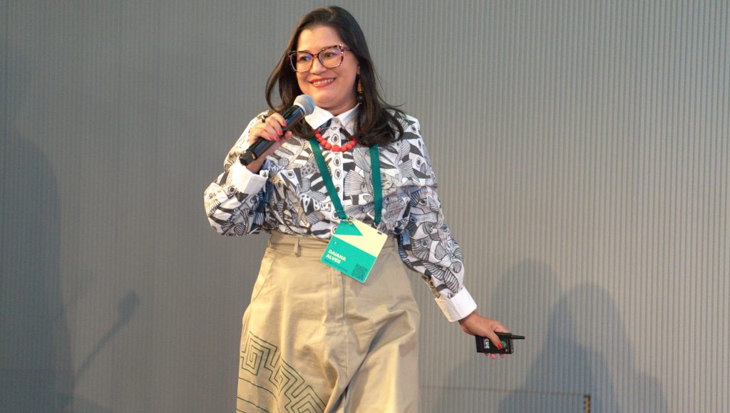 Fotografia de Daiana Alves. Ela caminha sorridente. Em uma mãe segura um microfone e na outra um pequeno aparelho eletrônico preto. Ela veste roupas sociais e usa um crachá na cor verde.