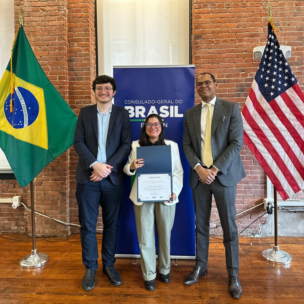 Fotografia posada de Daiana Alves, ao lado de dois homens. Todos se vestem de roupas sociais. Ela segura um certificado. Na lateral esquerda da foto há uma bandeira do Brasil e na lateral direita há uma bandeira dos Estados Unidos da América.