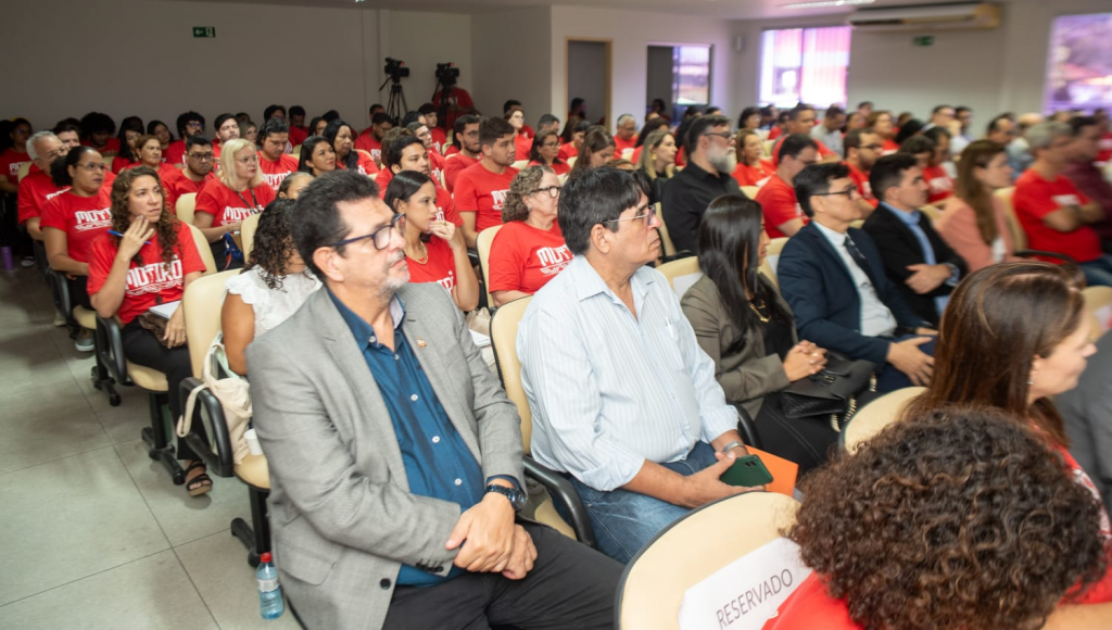 Fotografia da plateia de um auditório que está lotado. A grande maioria do público veste uma camisa vermelha.