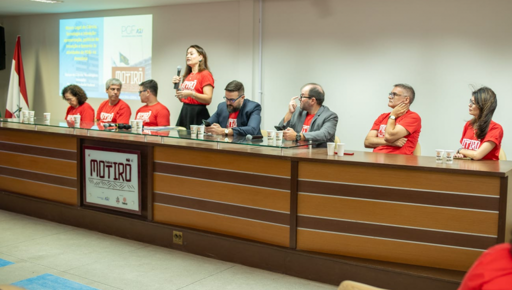 Fotografia registra a mesa de um evento. Nela estão oito pessoas e todas vestem uma camisa vermelha com a palavra "Motirõ".
