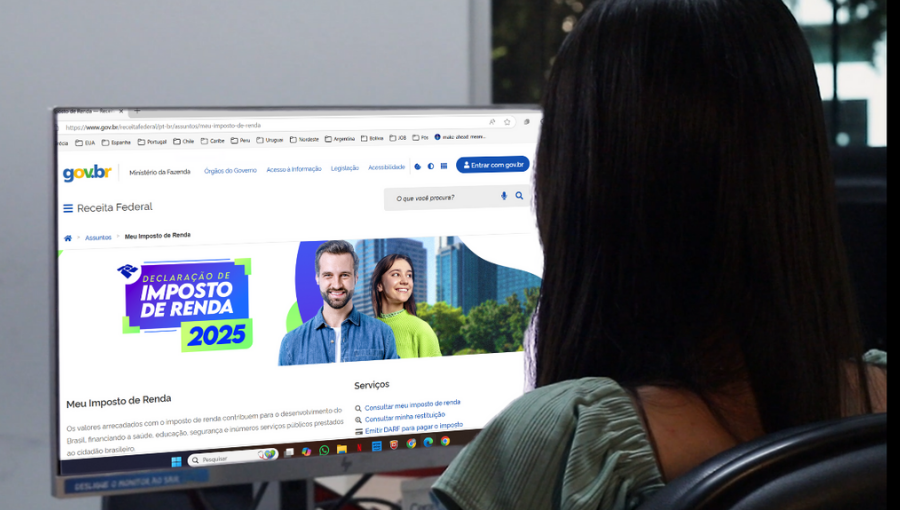 Fotografia registra uma pessoa, de cabelos compridos, de costa, em frente a uma tele de computador. Na tela, em destaque, a tela do site da Receita Federal, com uma imagem de um homem e uma mulher, seguido pelo texto: DECLARAÇÃO DE IMPOSTO DE RENDA 2025.