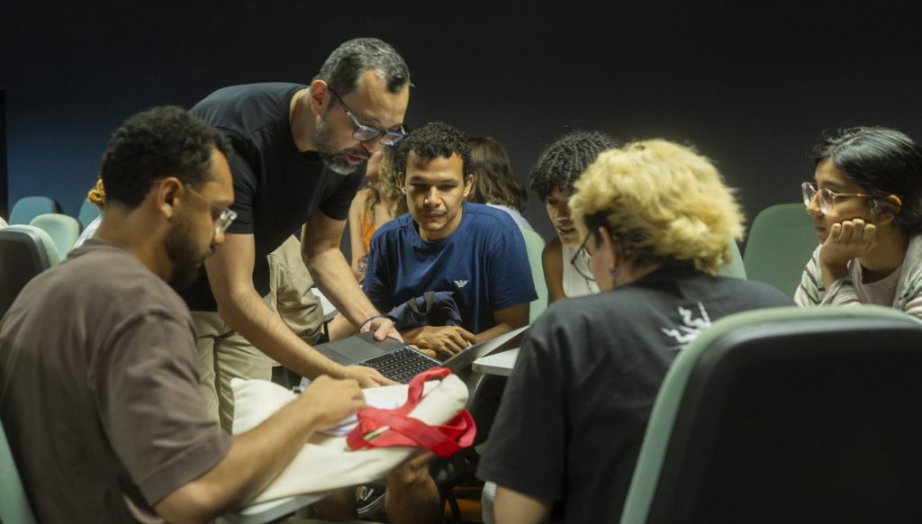 Fotografia de seis pessoas dispostas em um círculo. Uma pessoa, um homem, segura um notebook e mostra algo aos outros.