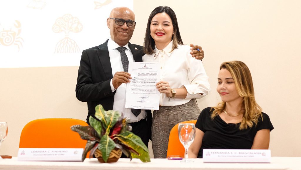 Fotografia posada da nova vice-coordenadora do Campus Soure, Leandra Cardoso Pinheiro, acompanhada pelo reitor da UFPA, Gilmar Pereira da Silva. Eles seguram um papel com o brasão da UFPA.