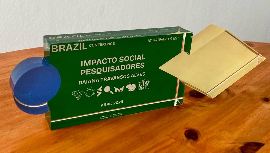 Fotografia de um troféu. Ele tem as cores verde, amarelo e azul. No troféu, há o seguinte texto: BRAZIL CONFERENCE - IMPACTO SOCIAL PESQUISADORES - DAIANA TRAVASSOS ALVES - 2025
